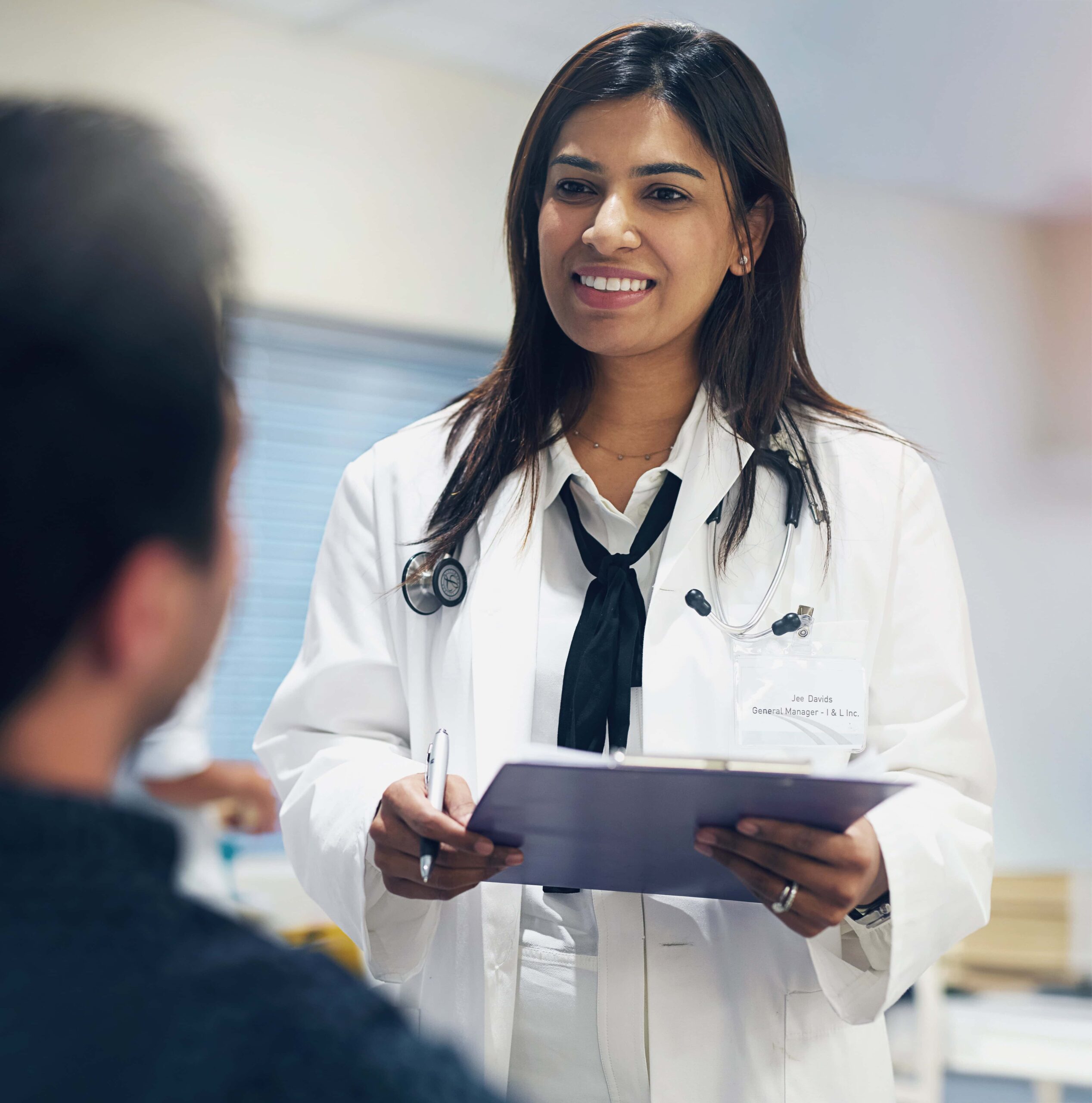 dedicated-to-her-patients-health-and-wellbeing-2025-04-06-07-47-35-utc female doctor standing smiling and holding clipboard while speaking to a male patient sitting down